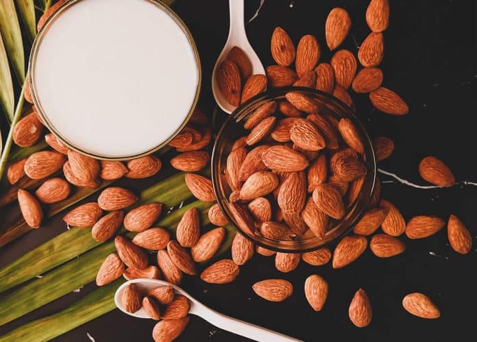 Top view of almonds scattered on a black surface with a glass of almond milk and spoons.