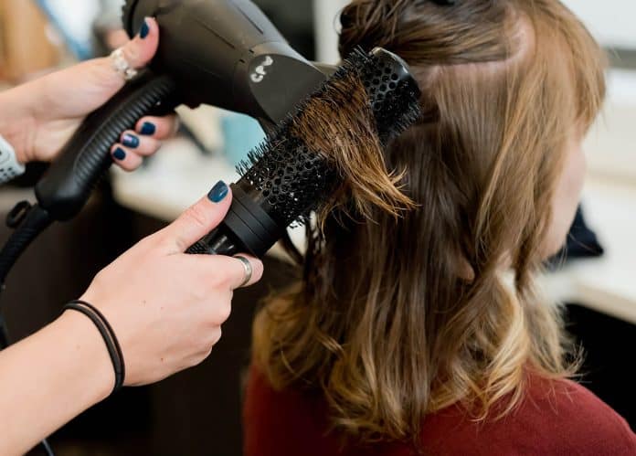 Close-up of a hairdresser blow-drying a customer's hair with a round brush in a salon setting.