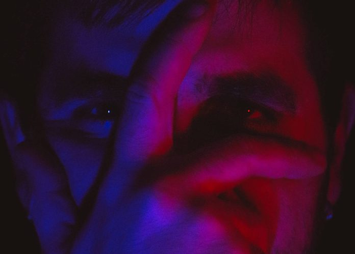 Close-up portrait of a person's face partially covered by their hand, illuminated by red and blue lighting for a dramatic effect.