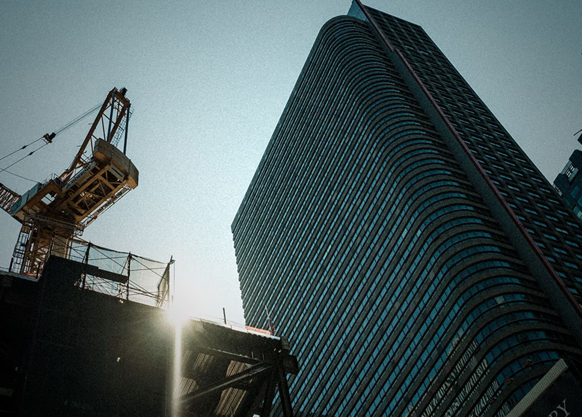 Photo of a construction crane beside a tall, curved glass building with sunlight filtering through.