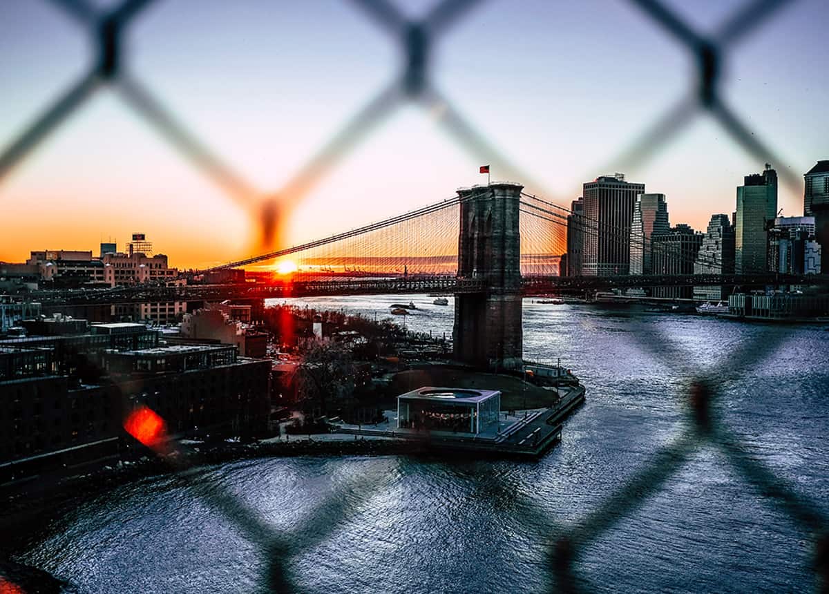 Sunset view of the Brooklyn Bridge and New York City skyline through a chain-link fence.