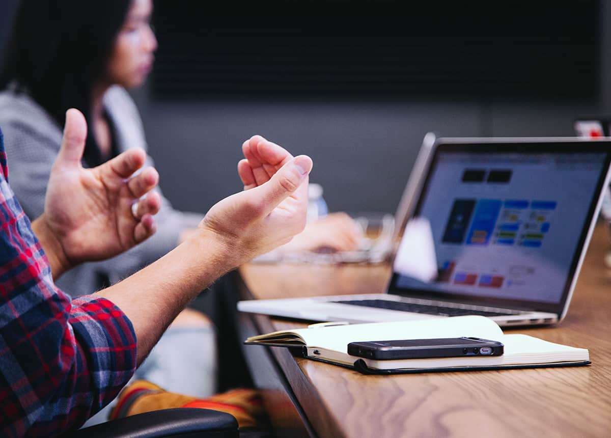 Photo of a person gesturing during a team discussion with a laptop and notes on the table.