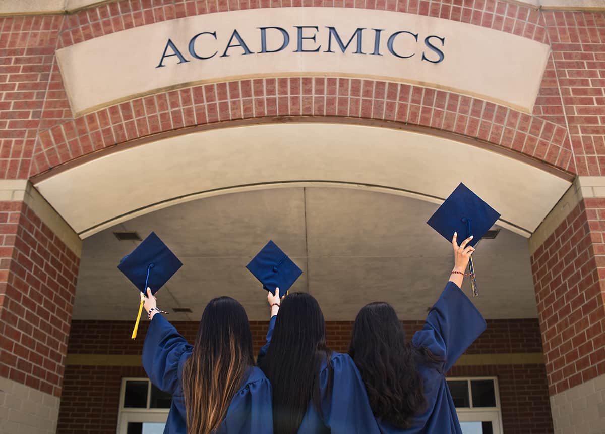 Three graduates in caps and gowns raising their caps in front of a building labeled 'ACADEMICS'.