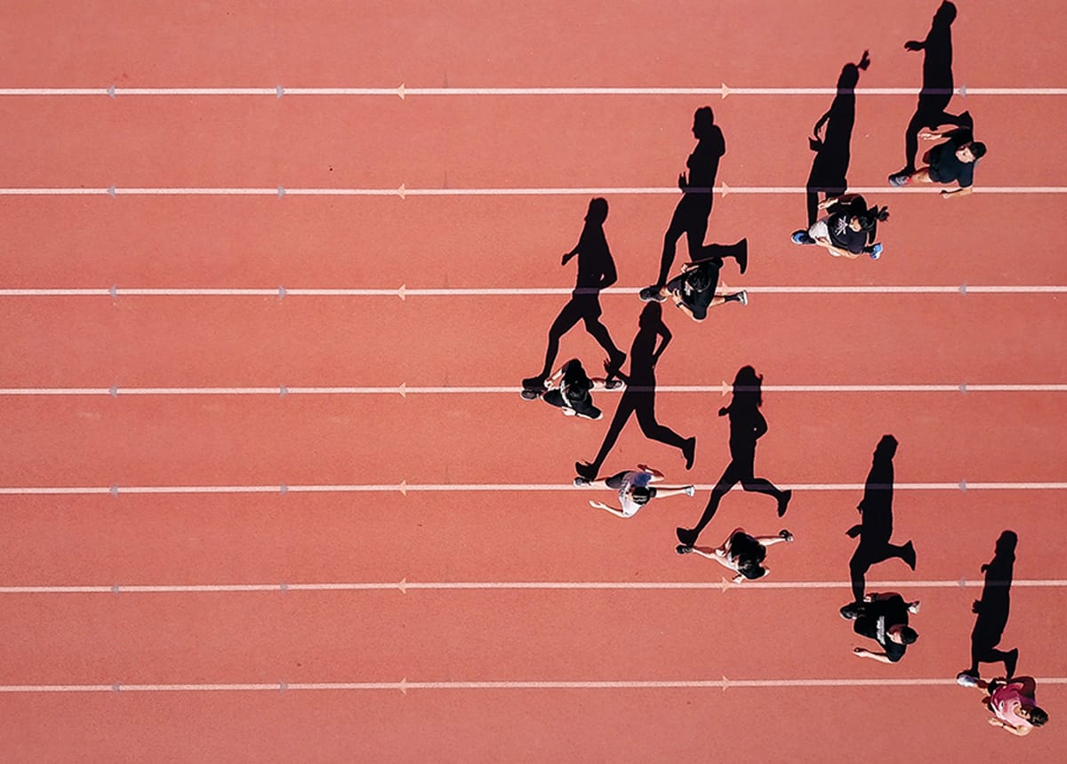 Overhead view of people running on a track, casting long shadows in the sunlight.